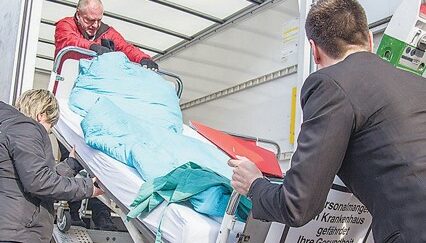 Foto: Die Linke @flickr Menschen heben ein Krankenhausbett, das voll Bezogen und mit Bettwäsche ausgestattet ist in einen Transporter.