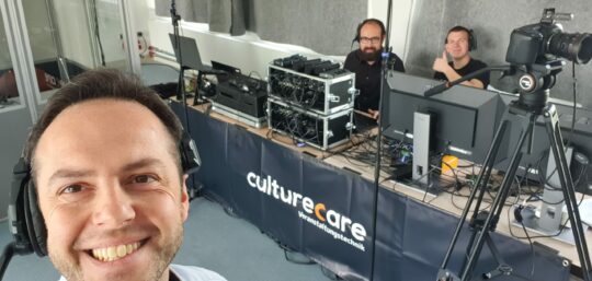 20230504_104034 Foto von Udo Nauber, der im Vordergrund steht und eine Headset trägt. Schräg hinter ihm ist diverse Veranstaltungstechnik auf einem Tisch mit einem Banner, das den Namen seiner Firma culturecare trägt, aufgebaut, unter anderem eine Kamera, Mischpulte und Computer. Hinter dem Tisch sitzen nicht namentlich bekannte Mitarbeiter der Firma. Beide tragen ebenso Headsets, und einer zeigt einen Daumen nach oben.