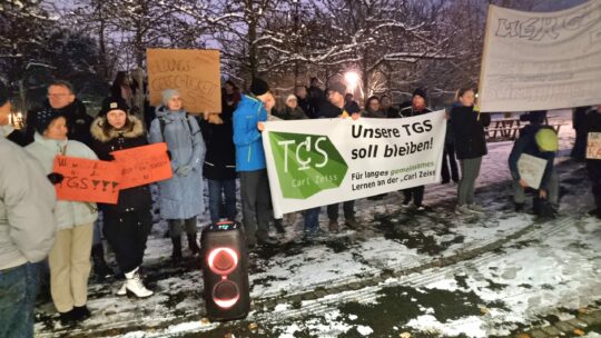 Unsere TGS Foto von Schülerinnen, Schülern und Eltern, welche Protestplakate und Transparente halten. Dabei liegt der Fokus zentral auf einem Transparent welches den Satz:"Unsere TGS soll bleiben", abbildet. Die Landschaft um die Menschenmenge ist schneebedeckt.