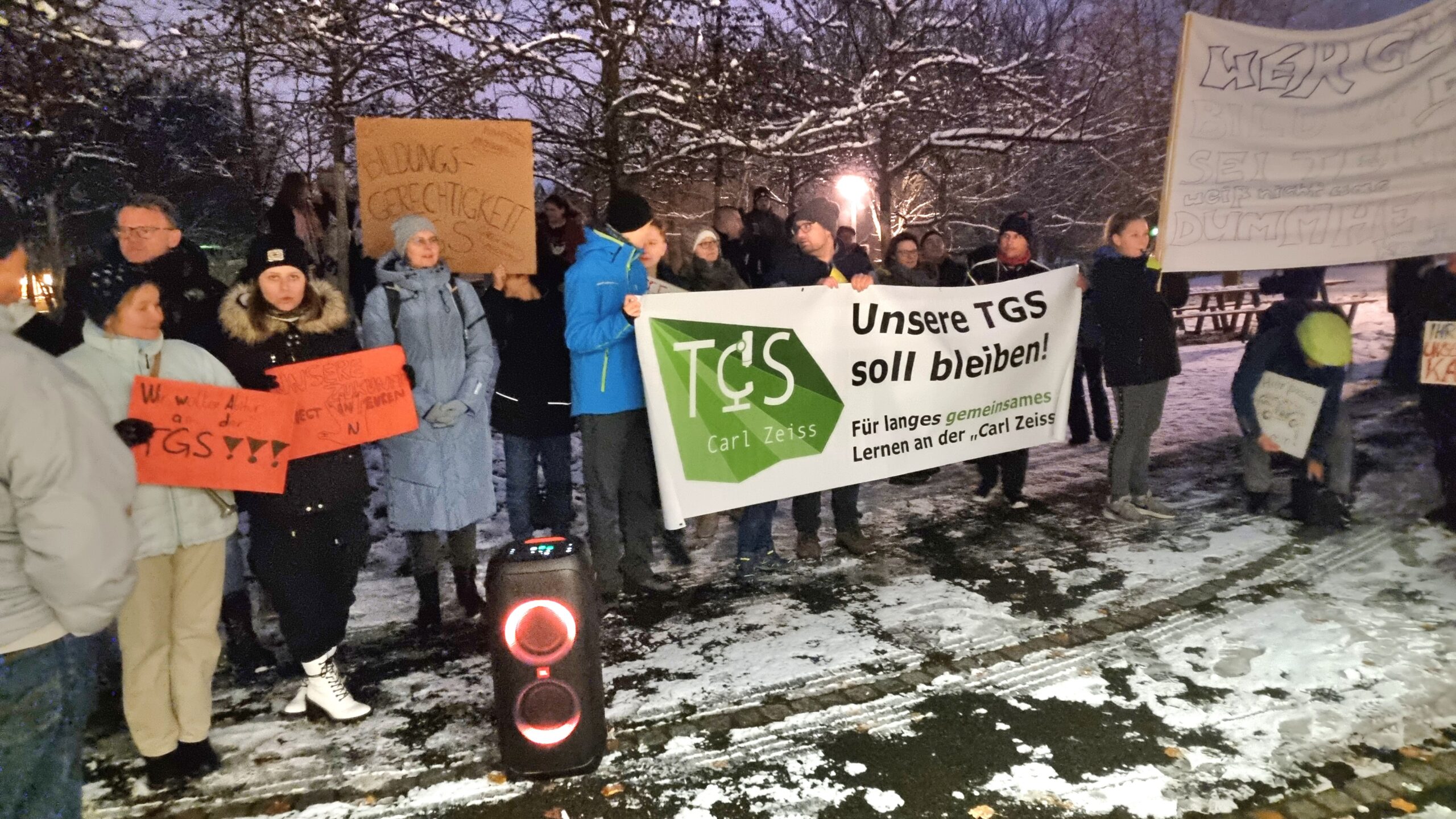 Unsere TGS Foto von Schülerinnen, Schülern und Eltern, welche Protestplakate und Transparente halten. Dabei liegt der Fokus zentral auf einem Transparent welches den Satz:"Unsere TGS soll bleiben", abbildet. Die Landschaft um die Menschenmenge ist schneebedeckt.