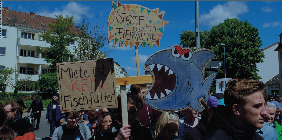 Screenshot 2024-07-12 103527 Eine Gruppe an Menschen nimmt an einen Protest teil. In der Bildmitte liegt der Fokus auf einem Demoplakat, welches Einen Hai zeigt welcher auf den Schriftzug:" Miete ist kein Fischfutter", zu schwimmt.