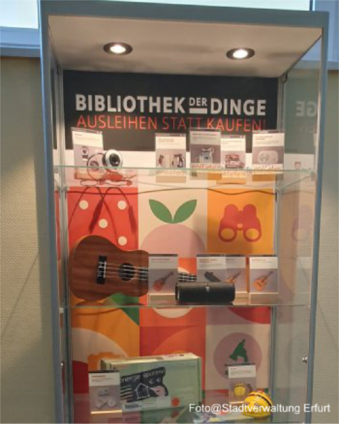 Vitrine der Bibliothek der Dinge in Erfurt Eine Galsvitrine der Bibliothek der Dinge in Erfurt, die auf unterschiedlichen Ebenen mit ausleihbaren Sachen bestückt sind. Darunter unter anderem eine Ukulele, eine Webcam, ein Spielzeug.