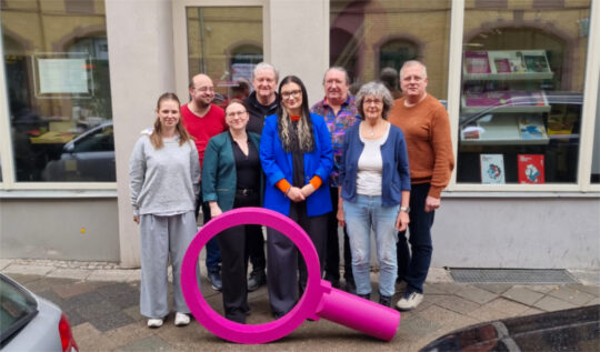 Aktueller Vorstand Die Thüringengestalter 2026 Eine Gruppe von Menschen steht eng beieinander auf einem Gehweg vor einem Gebäude mit großen Fenstern. Im Vordergrund befindet sich eine überdimensionale, beerenfarbige Lupe, die aufrecht auf dem Boden steht. Die Personen sind unterschiedlich gekleidet, darunter Jacken, Pullover und eine auffällige blaue Jacke. Im Hintergrund spiegeln sich geparkte Autos in den Fenstern. Die Szene zeigt den aktuellen Vorstand des Kommunalpolitischen Bildungsvereins Die Thüringengestalter.