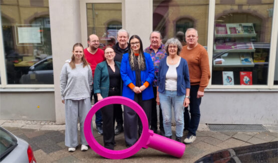 Aktueller Vorstand Die Thüringengestalter 2026 Eine Gruppe von Menschen steht eng beieinander auf einem Gehweg vor einem Gebäude mit großen Fenstern. Im Vordergrund befindet sich eine überdimensionale, beerenfarbige Lupe, die aufrecht auf dem Boden steht. Die Personen sind unterschiedlich gekleidet, darunter Jacken, Pullover und eine auffällige blaue Jacke. Im Hintergrund spiegeln sich geparkte Autos in den Fenstern. Die Szene zeigt den aktuellen Vorstand des Kommunalpolitischen Bildungsvereins Die Thüringengestalter.